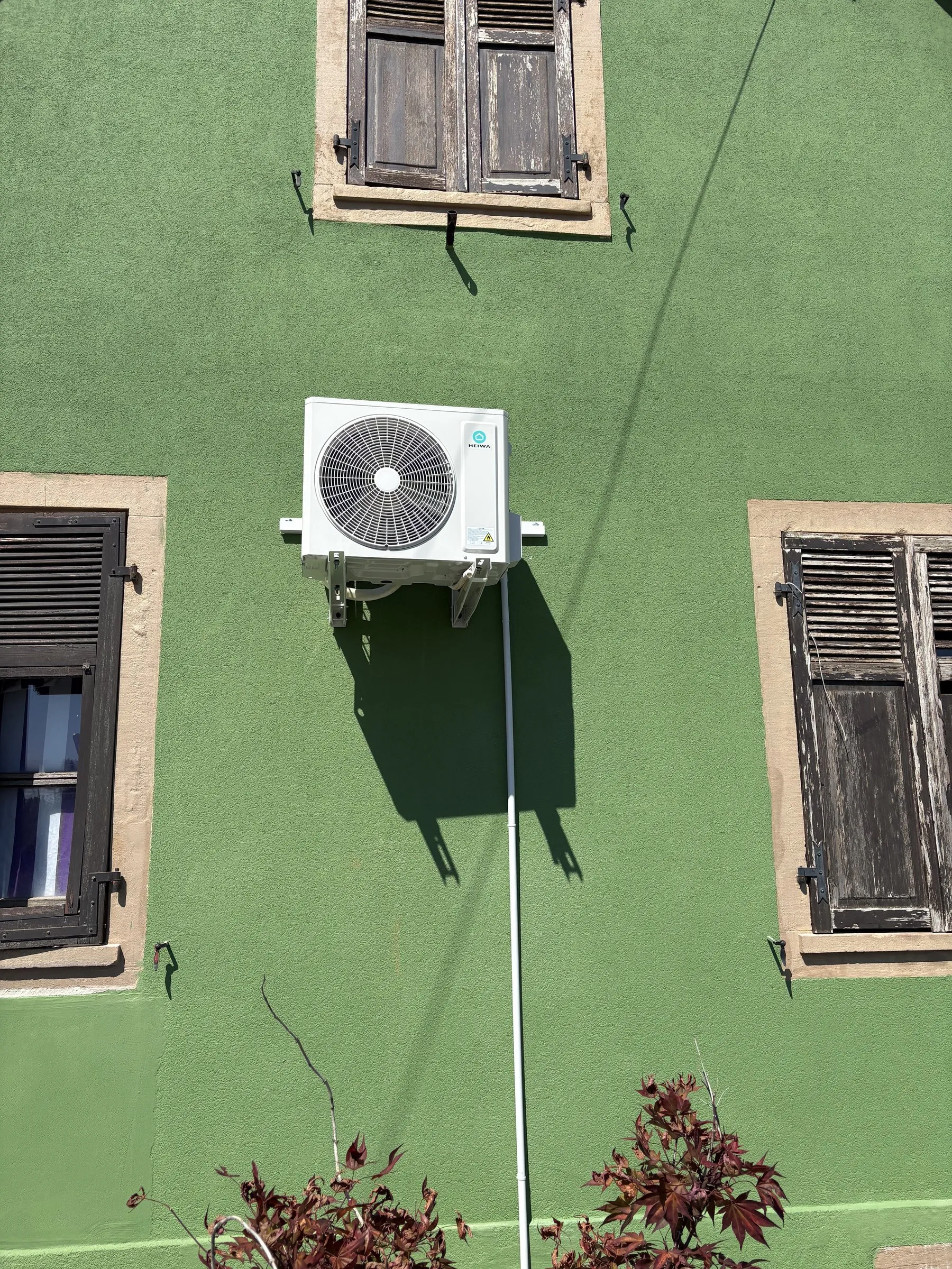 Unité extérieure de climatisation sur console, sur une façade verte entre deux fenêtres à volets