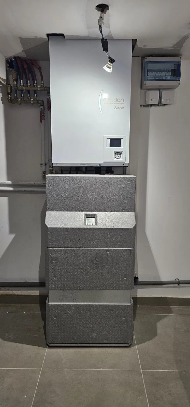 Module hydraulique de pompe à chaleur Mitsubishi Ecodan dans un local technique carrelé, à côté du tableau électrique