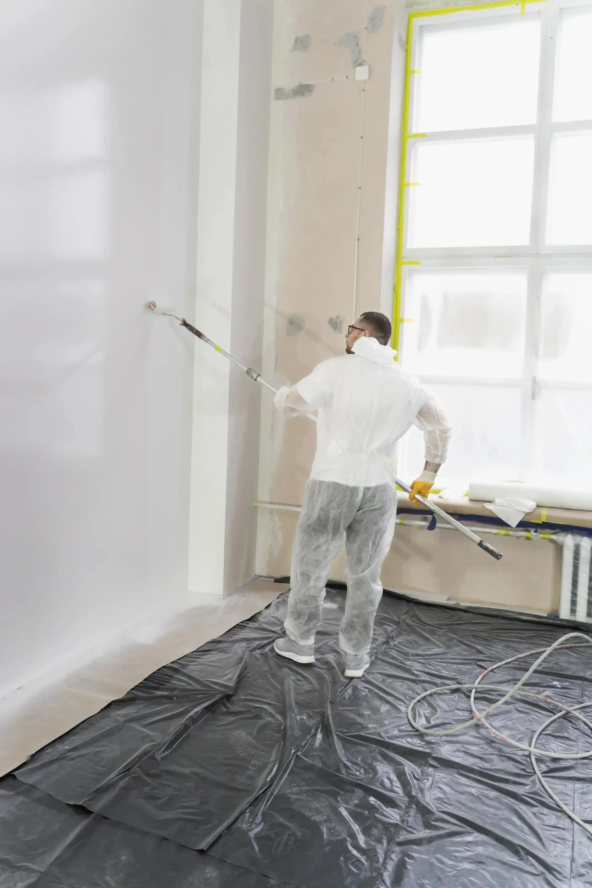 Peintre en combinaison appliquant la peinture au pistolet airless dans une pièce protégée